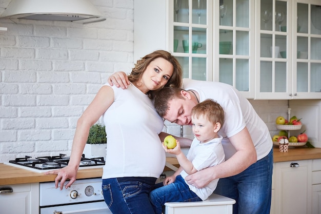 Eine dreiköpfige Familie mit einer schwangeren Frau und einem kleinen Sohn umarmt sich in einer großen weißen Küche.