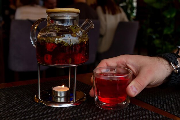 Foto eine durchsichtige glasteekanne mit rotem tee auf dem fußbrett und eine kerze steht auf dem tisch neben glas