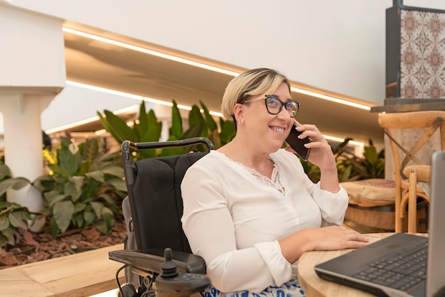 Eine elegante behinderte Frau sitzt im Rollstuhl im Café vor dem Laptop und telefoniert mit dem Handy