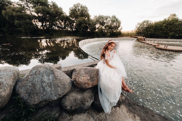 Eine elegante Braut in einem weißen Kleid, Handschuhen und nackten Füßen sitzt in der Nähe eines Wasserfalls im Park und genießt die Natur. Ein Modell in Hochzeitskleid und Handschuhen in einem Naturpark. Weißrussland