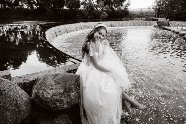 Eine elegante Braut in einem weißen Kleid, Handschuhen und nackten Füßen sitzt in der Nähe eines Wasserfalls im Park und genießt die Natur. Ein Modell in Hochzeitskleid und Handschuhen in einem Naturpark. Weißrussland