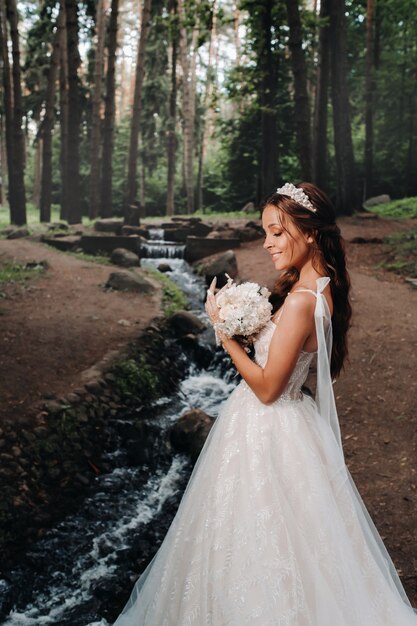 Eine elegante Braut in einem weißen Kleid und Handschuhen mit einem Blumenstrauß steht an einem Bach im Wald und genießt die Natur. Ein Modell in einem Hochzeitskleid und Handschuhen in einem Naturpark. Weißrussland