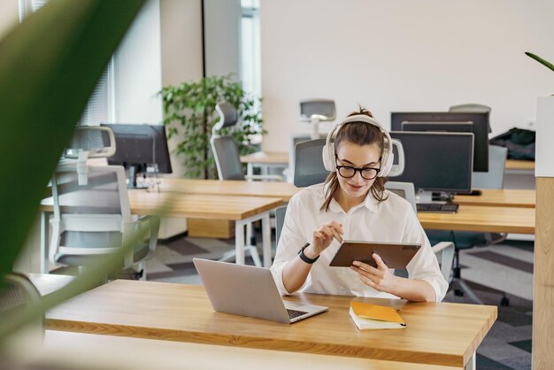 Eine erfahrene Frau mit Kopfhörern benutzt ein Tablet mit einem scharfen Fokus, das in einer geräumigen Wohnung in der Nähe eines Laptops sitzt