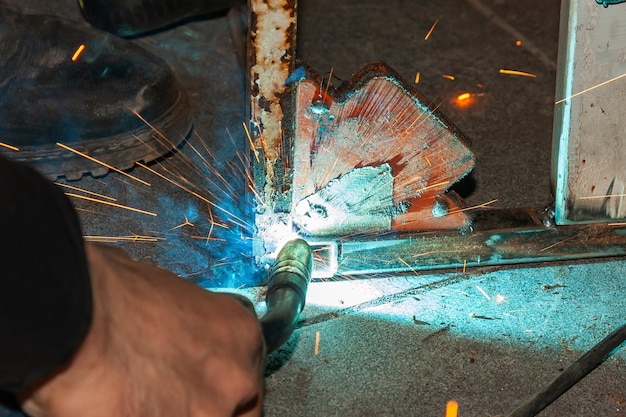 Eine erfahrene Person führt Arbeiten mit einer Schweißmaschine durch, repariert Metallteile und entfernt blauen Rauch sowie gelbe Funken und Blitze in der industriellen Produktionswerkstatt