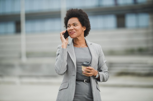 Eine erfolgreiche schwarze Geschäftsfrau, die eine App auf einem Smartphone verwendet, während sie eine Kaffeepause vor einem Firmengebäude macht.
