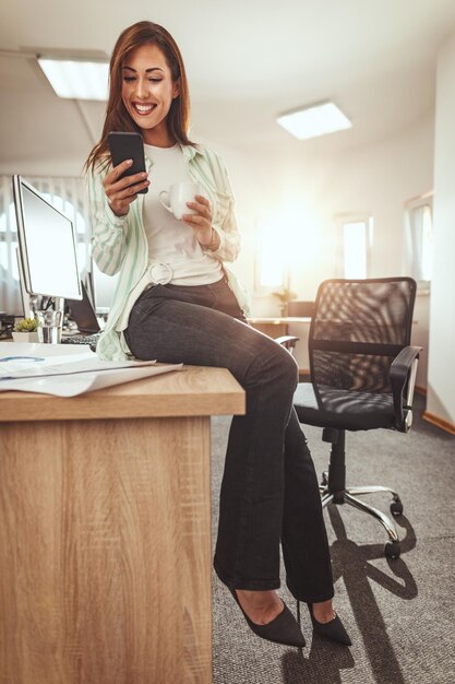 Eine erfolgreiche Unternehmerin sitzt am Bürotisch und schreibt Nachrichten auf ein Smartphone und trinkt Kaffee im Büro.