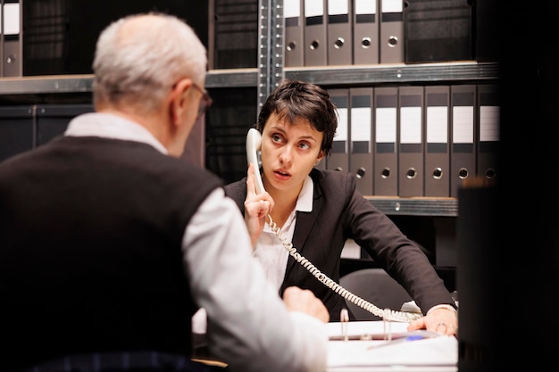 Foto eine ermittlerin spricht am festnetztelefon mit einem ferninspektor und bespricht den fall einer vermissten person. älterer polizist analysiert verbrechensbeweise und macht überstunden im archivraum