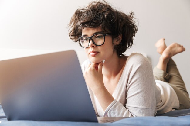 Eine ernsthafte junge Frau liegt drinnen mit einem Laptop auf dem Bett.