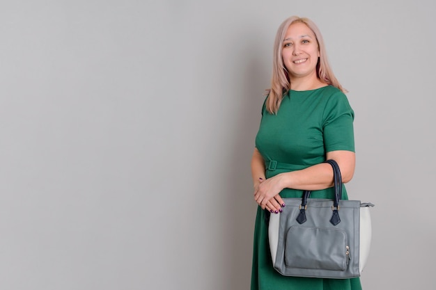 Eine erwachsene blonde Frau in einem grünen Kleid mit einer Handtasche steht auf einem grauen Hintergrund, Exemplar