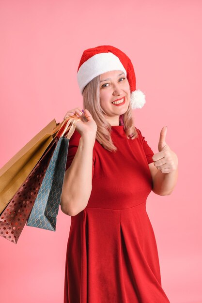 Eine erwachsene Frau in einer Weihnachtsmütze mit Einkaufstüten zeigt ihre Daumen nach oben und posiert auf einem rosa Hintergrund