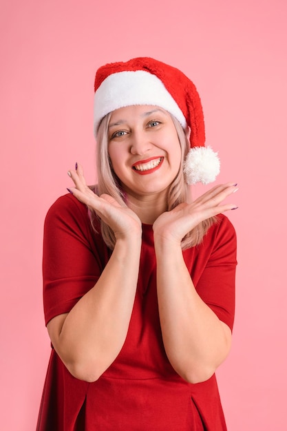 Eine erwachsene Frau in einer Weihnachtsmütze stützt ihr Kinn mit den Händen auf einem rosa Hintergrund, vertikales Foto