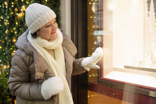 Foto eine erwachsene frau in winterkleidung betrachtet ein weihnachtsschaufenster und lächelt.