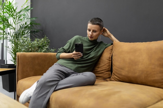 Eine erwachsene Frau mit kurzem Haarschnitt sitzt zu Hause auf der Couch mit einem Handy in der Hand