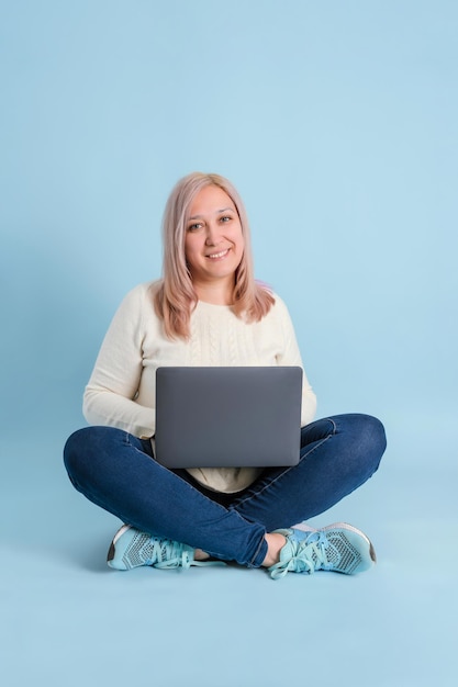 Eine erwachsene Freiberuflerin mit einem Laptop auf dem Schoß sitzt auf blauem Hintergrund