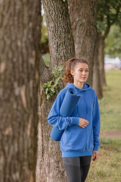 Eine europäerin in einem blauen Hoodie mit blauer Yogamatte wählt einen Ort für Sport und Meditation