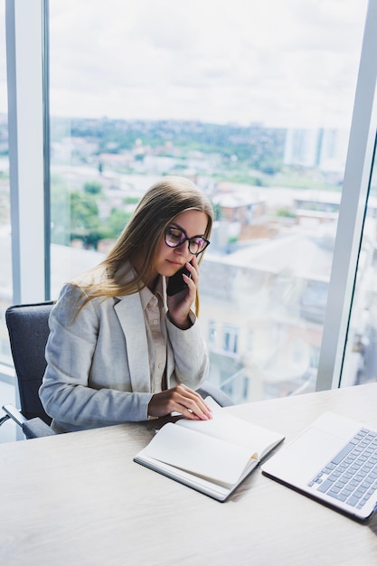 Eine fairhaarige, fröhliche Europäerin mit Brille in stilvoller Freizeitkleidung sitzt an einem Tisch mit einem Laptop, der Papierkram erledigt und telefoniert Geschäftsfrau am Arbeitsplatz im Büro