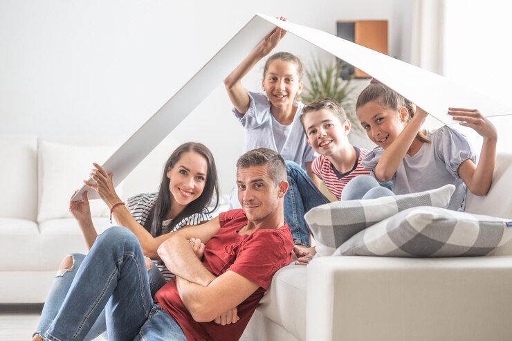 Eine Familie mit drei Kindern sitzt glücklich unter einem symbolischen Dach zu Hause auf und um das Sofa herum