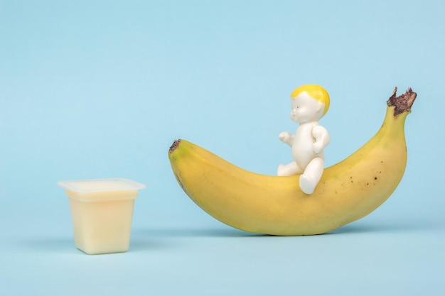Eine Figur eines Babys, das auf einer Banane reitet und eine Tasse Bananendessert auf blauem Hintergrund. Das Konzept der gesunden Babynahrung.