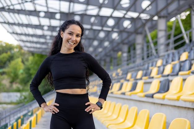 Eine fitte und glückliche Frau in Sportkleidung steht zuversichtlich in einem Stadion und strahlt ein Lächeln aus.