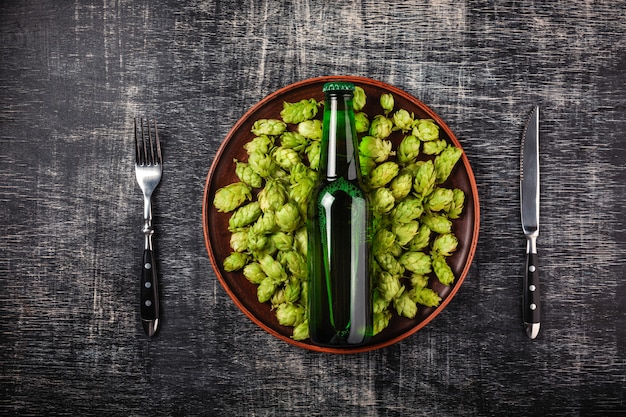 Eine Flasche Bier auf einem grünen frischen Hopfen in einer Platte mit Messer und Gabel gegen den Hintergrund
