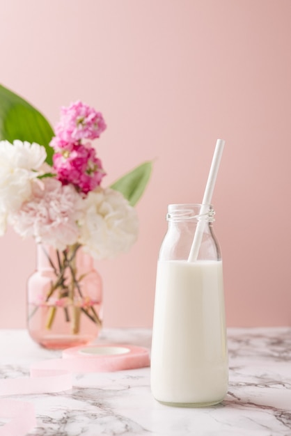 Eine Flasche Milch auf Marmortisch auf rosa Hintergrund mit rosa und weißem Blumenstrauß, heller und pastellfarbener Getränkezusammensetzung vertikal composition