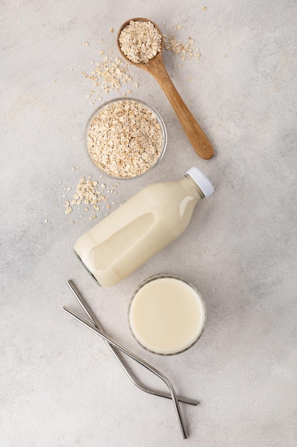 Eine Flasche und ein Glas pflanzliche Hafermilch mit einer Zutat in einer Schüssel mit wiederverwendbaren Strohhalmen auf hellem Hintergrund