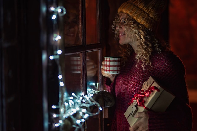 Eine Frau allein zu Hause trinkt Tee in der Weihnachtsferienzeit, die ein Geschenk hält und bei Nacht aus dem Fenster schaut Glückliche Menschen und Dezemberfeier Silvester und Innenhausatmosphäre