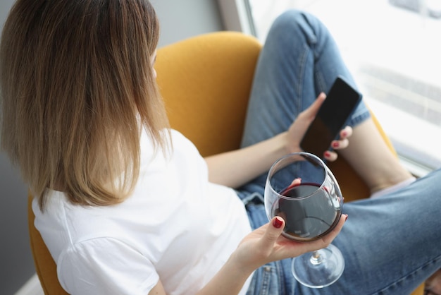 Eine Frau am Fenster hält ein Glas Rotwein und ein Smartphone, Draufsicht, Nahaufnahme. Einsamkeit, Stressabbau, Selbstisolation