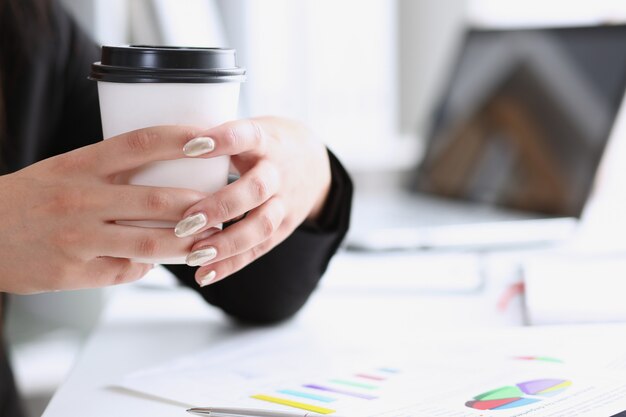 Eine Frau an einem Arbeitsplatz im Büro hält eine weiße Tasse mit Kaffee in den Händen