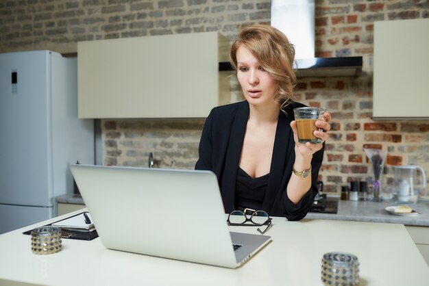 Eine Frau arbeitet in ihrer Küche an einem Laptop. Eine Dame hält ein Glas Kaffee in der Hand und bereitet sich auf einen Vortrag bei einem Videoanruf vor.