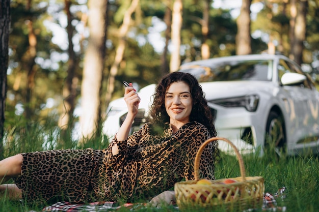 Eine Frau bei einem Picknick in der Natur, im Hintergrund wird ein Elektroauto aufgeladen