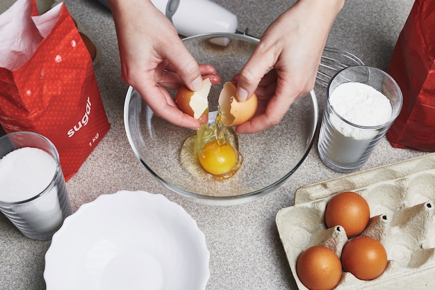 Eine Frau bricht ein Ei in eine transparente Schüssel. Zutaten zum Backen von Kuchen auf einem Tisch.