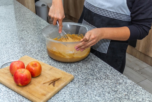 Eine Frau, deren Gesicht nicht zu sehen ist, mischt den Teig für einen Kuchen neben einer Gruppe Äpfel in der Küche ihres Hauses. Verfahren zur Herstellung von Apfelkuchen