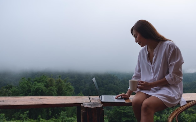 Eine Frau, die an einem Laptop-Computer benutzt und arbeitet, während sie an einem nebligen Tag auf dem Balkon mit einem wunderschönen Blick auf die Natur sitzt