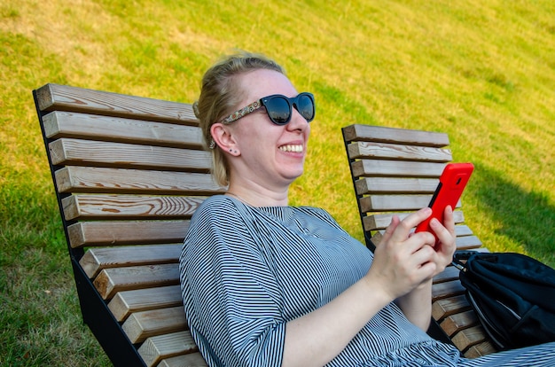 Eine Frau, die an einem Sommertag in einem Park sitzt, kauft online ein oder kommuniziert in Instant Messenger von ihrem Smartphone aus