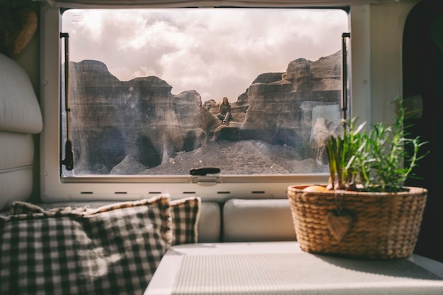 Eine Frau, die auf den Felsen sitzt, blickt von innen auf einen modernen Wohnmobil durch das Fenster