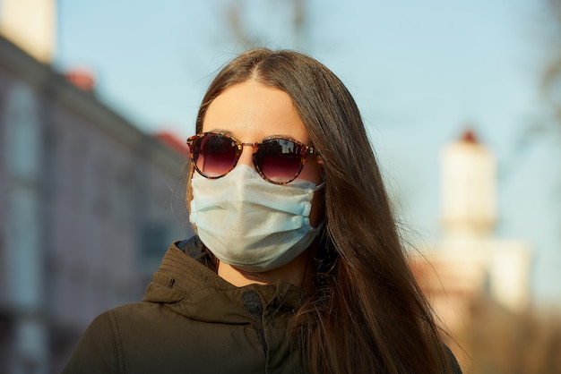 Eine Frau, die eine medizinische Gesichtsmaske trägt, um die Ausbreitung des Coronavirus auf einer Stadtstraße bei Sonnenuntergang zu vermeiden.