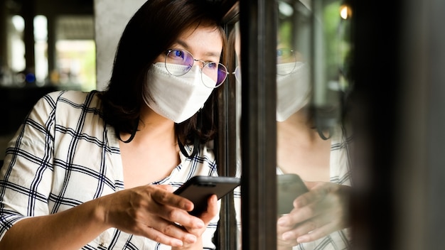 Eine Frau, die eine medizinische Maske mit einem Smartphone trägt, schaut aus dem Fenster, sie langweilt sich und nutzt während des Coronavirus-Ausbruchs zu Hause soziale Medien.