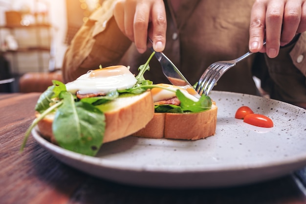 Eine Frau, die Frühstückssandwich mit Eiern, Speck und saurer Sahne durch Messer und Löffel in einem Teller auf Holztisch isst