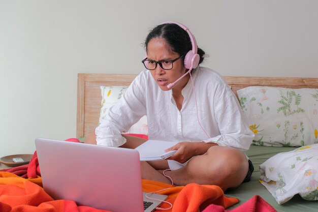 Foto eine frau, die im bett sitzt und auf ihren laptop schaut, ein notizbuch in ihrer hand, ärgert verwirrten gesichtsausdruck