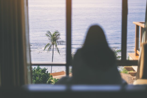 Eine Frau, die im Bett sitzt und schönen Meerblick durch das Fenster betrachtet, nachdem sie am Morgen aufwacht