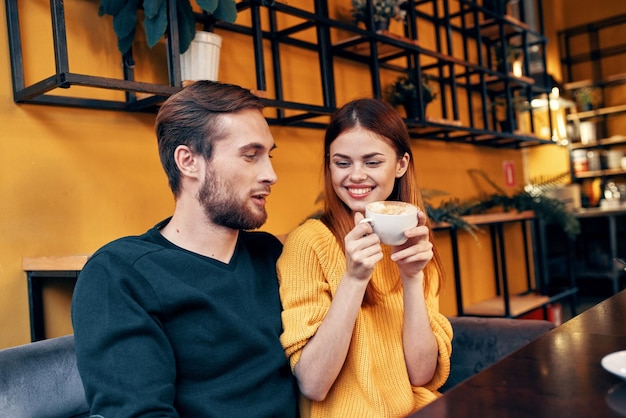 Eine Frau, die in eine Tasse Kaffee verliebt ist, und ein glücklicher Mann an einem Tisch in einem Café-Interieur mit Freunden