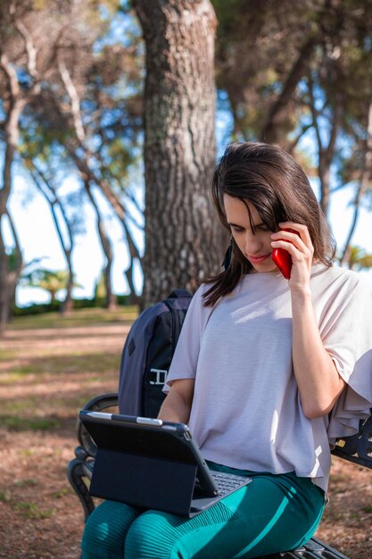 Eine Frau, die in einem Park mit einem Laptop arbeitet und auf dem Smartphone spricht Digitaler Nomade für Telearbeit