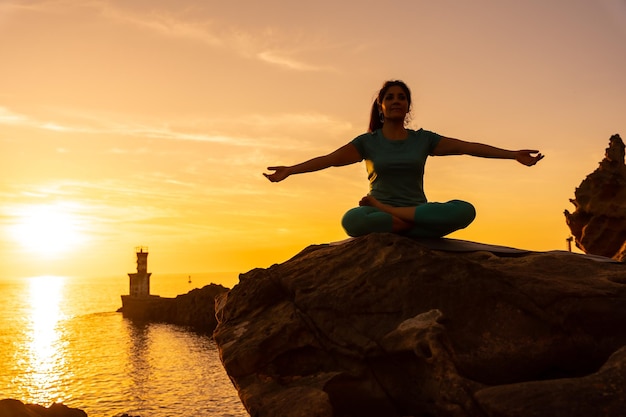 Eine Frau, die Meditations- und Yoga-Übungen bei Sonnenuntergang neben einem Leuchtturm im Meer macht gesundes und naturistisches Leben im Freien pilates ardha padmasana