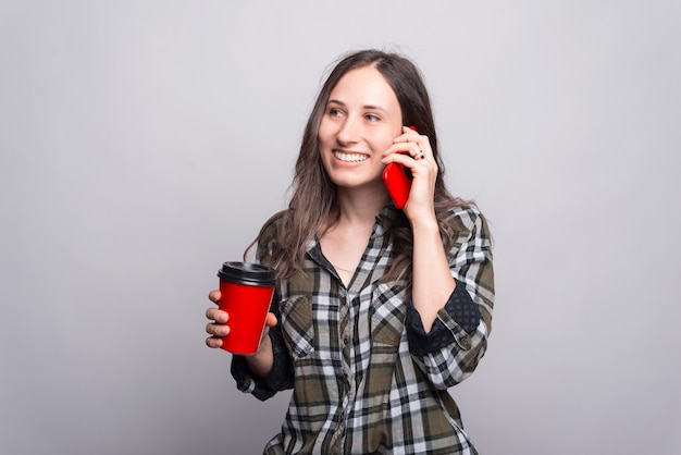 Eine Frau, die mit ihrem Telefon spricht, lächelt und hält eine Tasse mit heißem zu trinken nahe einer grauen Wand