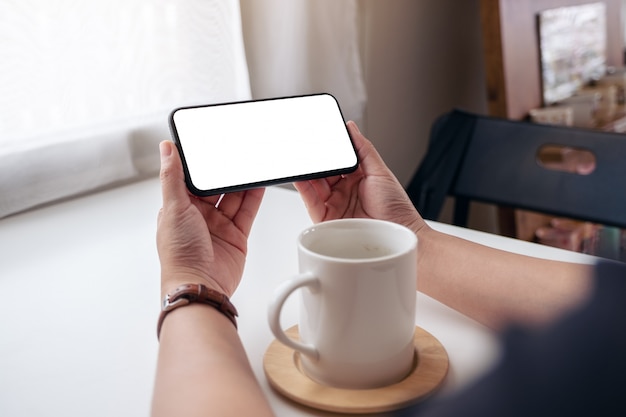 eine Frau, die schwarzes Handy mit leerem Desktop-Bildschirm mit Kaffeetasse auf dem Tisch hält