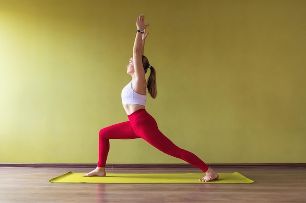 Eine Frau, die Yoga praktiziert, führt die Virabhadrasana-Übung Kriegerpose im Studio in Sportkleidung in der Nähe der Wand aus
