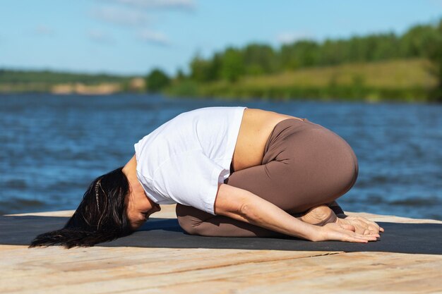 Eine Frau führt in Sportbekleidung am Ufer eines Sees eine Balasana-Übung durch. Kinderpose trainiert