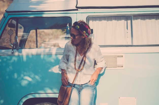 Eine Frau gegen den blauen klassischen Van reisen und allein Hippie-Lifestyle-Freizeitaktivitäten im Freien genießen Konzept des Fahrzeugurlaubs und des Vanlife Lässige, trendige weibliche Menschen Kleidung im Retro-Stil und Wohnmobil