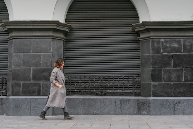 Eine Frau geht eine alte Straße entlang, schaut auf den Boden und hat ihre Hände in ihrem langen Mantel vor dem Hintergrund alter Graustufenarchitektur. Platz kopieren